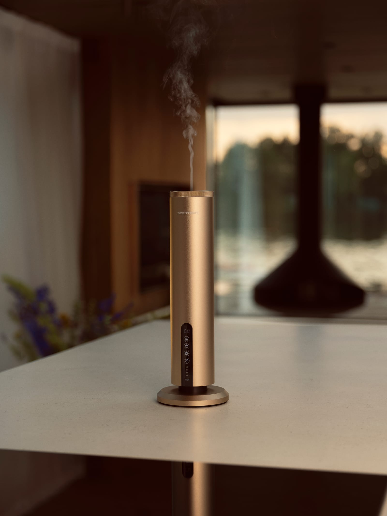 Scentech Smart Aroma Diffuser Pro releasing mist on a white table, a fireplace and the lake blurred behind it